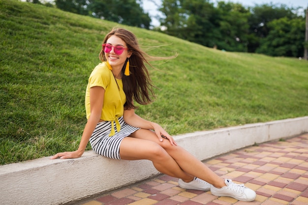 Young attractive stylish smiling woman having fun in city park, positive, emotional, wearing yellow top, striped mini skirt, pink sunglasses, white sneakers, summer style fashion trend, long legs