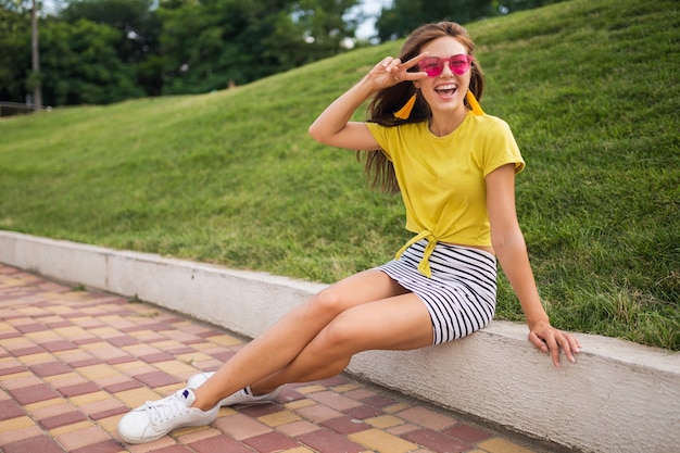 Young attractive stylish smiling woman having fun in city park, positive, emotional, wearing yellow top, striped mini skirt, pink sunglasses, white sneakers, summer style fashion trend, peace sign