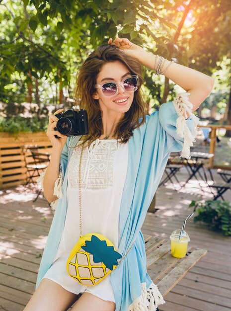 Young attractive stylish woman in park, street style, summer fashion trend, blue cape, white boho dress, accessories, holding vintage photo camera, smiling, happy emotion, enjoying warm sunny day