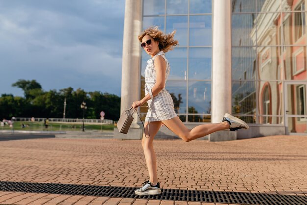 Young attractive stylish woman running jumping funny in sneakers in city street in summer fashion style white dress wearing sunglasses and handbag