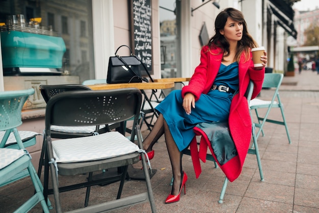 Young attractive stylish woman sitting in city street cafe in red coat, autumn style fashion trend, drinking coffee, wearing blue dress, high heeled shoes, legs in black net stockings, elegant lady