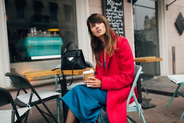 Young attractive stylish woman sitting in city street cafe in red coat drinking coffee wearing blue dress