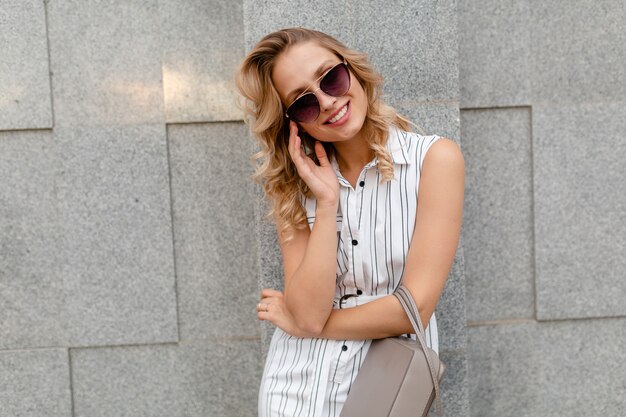 Young attractive stylish woman with blond curly hairstyle walking in city street in summer fashion style white striped dress wearing sunglasses holding purse