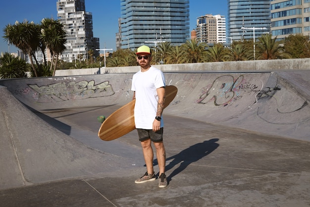 Young attractive tattooed skater in trucker cap stands in unlabeled blank white t-shitrt with his wooden longboard in hand in center of skatepark, urban landscape behind