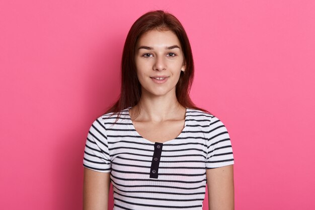 Молодая привлекательная девочка-подросток в полосатой повседневной футболке, позирует у розовой стены, с очаровательной улыбкой, выглядит счастливой.