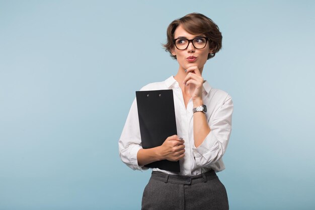 Young attractive thoughtful woman with dark short hair in shirt and eyeglasses holding black folder in hand while dreamily looking aside over blue background