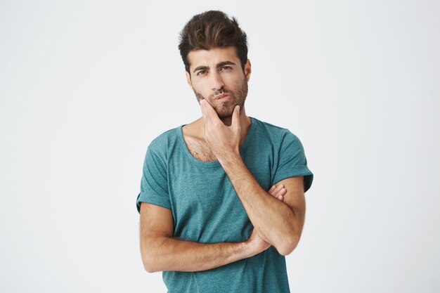 Young attractive trendy hispanic student in blue t-shirt, touching face with hand thoughtfully looking at university schedule trying find out free time for work.