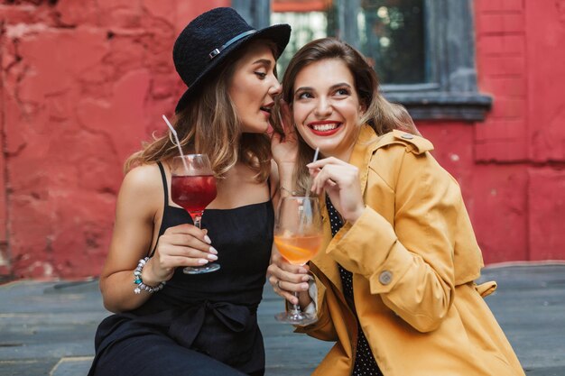 Young attractive woman in black dress and hat thoughtfully talking gossips to pretty smiling lady holding cocktails in hands while spending time together in old courtyard of cafe