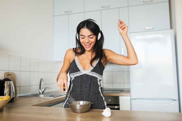 Young attractive woman cooking scrambled eggs in kitchen in morning, smiling, happy positive housewife, healthy lifestyle, listening to music on headphones, laughing, having fun, dancing, singing