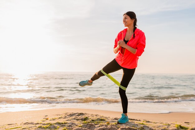 Young attractive woman doing sport exercises in morning sunrise on sea beach, healthy lifestyle, listening to music on earphones, wearing pink windbreaker jacket, making stretching in rubber band