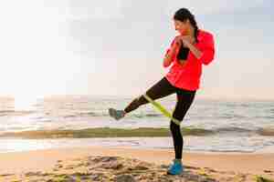 Free photo young attractive woman doing sport exercises in morning sunrise on sea beach, healthy lifestyle, listening to music on earphones, wearing pink windbreaker jacket, making stretching in rubber band