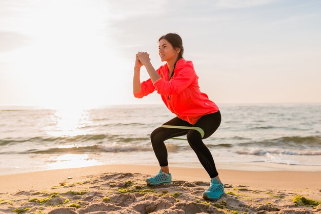 Young attractive woman doing sport exercises in morning sunrise on sea beach, healthy lifestyle, listening to music on earphones, wearing pink windbreaker jacket, making stretching in rubber band