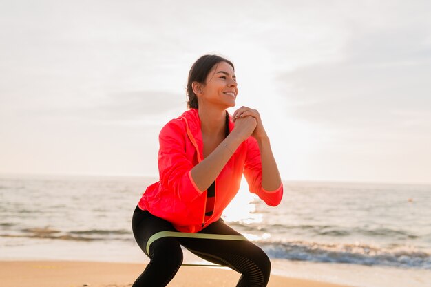 Young attractive woman doing sport exercises in morning sunrise on sea beach, healthy lifestyle, listening to music on earphones, wearing pink windbreaker jacket, making stretching in rubber band