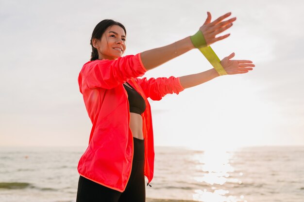 Молодая привлекательная женщина делает спортивные упражнения в утреннем восходе солнца на морском пляже, здоровый образ жизни, слушает музыку в наушниках, носит розовую куртку-ветровку, делает растяжку в резинке