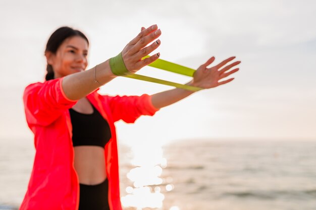 Young attractive woman doing sport exercises in morning sunrise on sea beach, healthy lifestyle, listening to music on earphones, wearing pink windbreaker jacket, making stretching in rubber band
