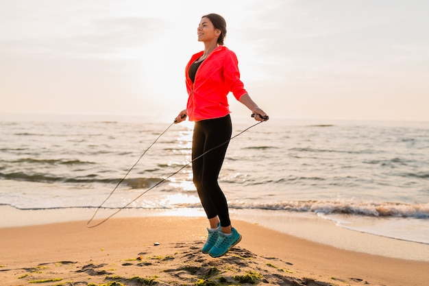 Young attractive woman doing sport exercises in morning sunrise on sea beach in sports wear, healthy lifestyle, listening to music on earphones, wearing pink windbreaker jacket, jumping in jump rope
