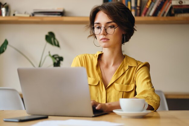 안경을 쓴 젊은 매력적인 여성이 현대 사무실에서 커피 한 잔과 함께 노트북 작업을 꿈꾸며 꿈꾸고 있습니다.