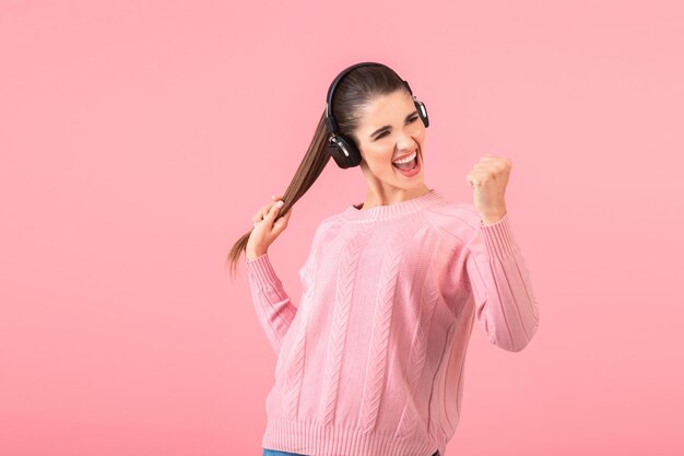 Молодая привлекательная женщина слушает музыку в беспроводных наушниках в розовом свитере, улыбаясь счастливым позитивным настроением, позируя на розовом фоне, изолированное смешное эмоциональное выражение лица