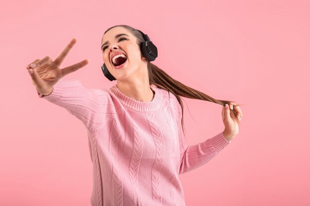 분홍색 배경에 포즈 행복 긍정적 인 분위기 미소 분홍색 스웨터를 입고 무선 헤드폰에서 음악을 듣고 젊은 매력적인 여자