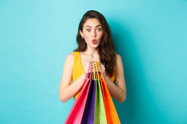 Young attractive woman looks intrigued at promo deal, holding shopping bags with goods, standing over blue background. Copy space