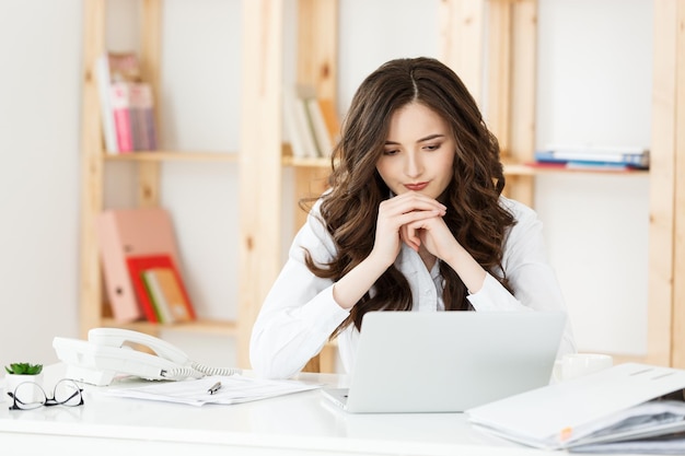 Giovane donna attraente a una scrivania da ufficio moderna, lavorando con il computer portatile e pensando a qualcosa