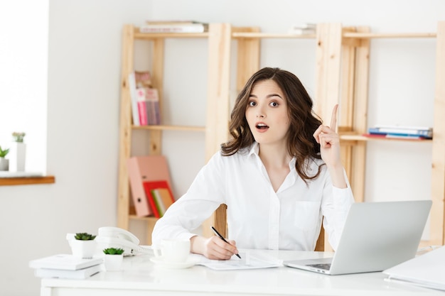 Giovane donna attraente a una scrivania da ufficio moderna, lavorando con il computer portatile e pensando a qualcosa