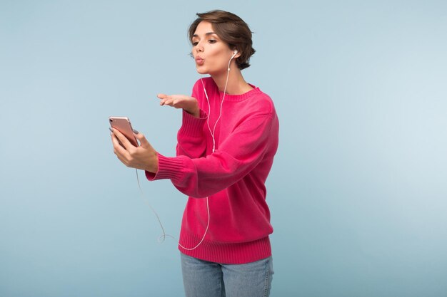 Young attractive woman in pink sweater and earphones dreamily sending air kiss while holding cellphone in hand over blue background