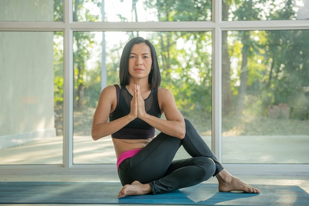 Молодая привлекательная женщина, практикующая йогу дома, Ардха Матсиендрасана, позирует с намастэ, тренируется, носит спортивную одежду, брюки и топ, закрытый полный рост, студия йоги.