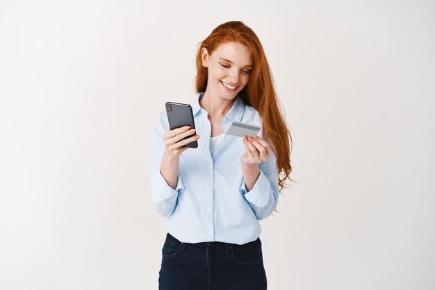 인터넷 흰색 배경에서 스마트폰과 플라스틱 신용 카드로 온라인 쇼핑을 하는 젊은 매력적인 여성