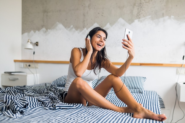 Young attractive woman sitting on bed in pajamas, smiling in bedroom, happy emotion, wake up in morning, laughing, sexy, skinny, listening to music on headphones, using smartphone, taking selfie photo