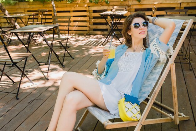 Young attractive woman sitting in deck chair in summer fashion outfit, white dress, blue cape, sunglasses, smiling, drinking lemonade, stylish accessories, relaxing on vacation