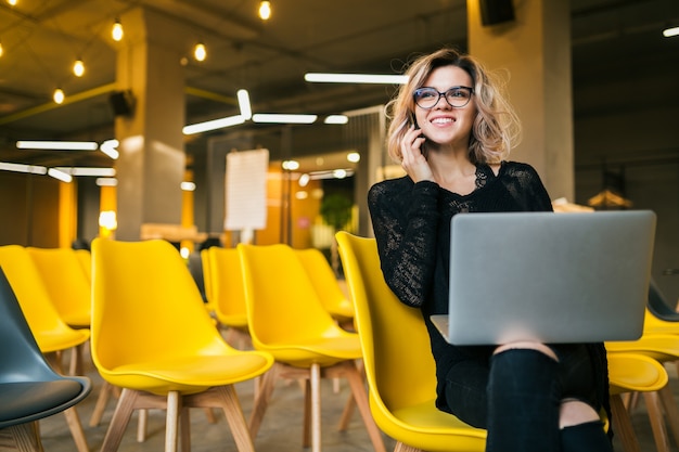 Young attractive woman sitting in lecture hall, working on laptop, wearing glasses, many yellow chairs, student education online, freelancer, smiling, talking on smartphone, looking forward, startup