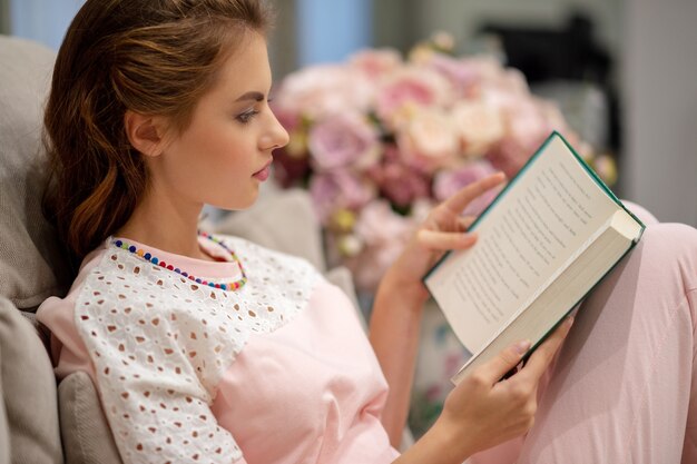 Молодая привлекательная женщина, сидящая на софе, читая книгу, наслаждается отдыхом.