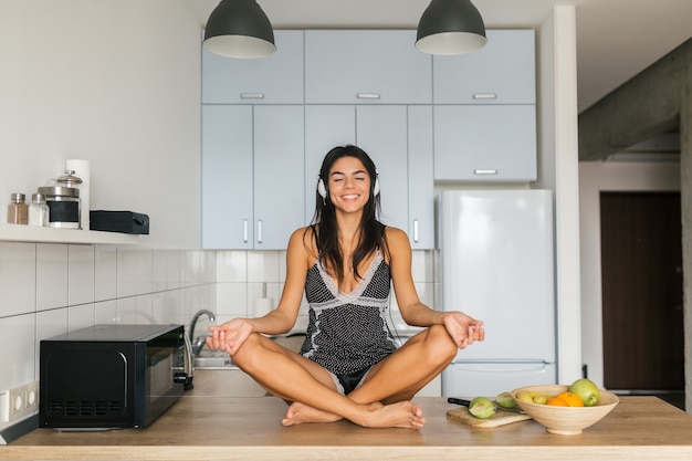 Foto gratuita giovane donna attraente che si siede asana yoga posa in cucina al mattino, maditating, sorridente, felice, positivo, stile di vita sano, ascolto di musica in cuffia, relax, armonia