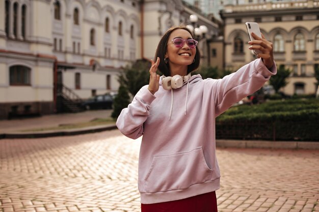 Young attractive woman in stylish hoodie and pink sunglasses smiles sincerely holds phone and takes selfie outside