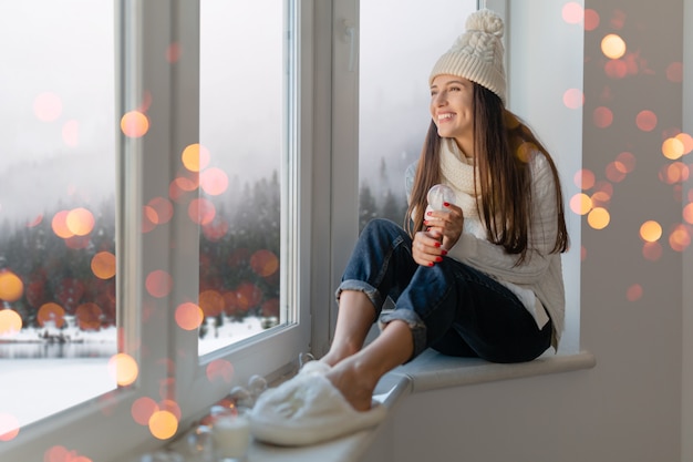 スタイリッシュな白いニットのセーター、スカーフと帽子の若い魅力的な女性は、ガラスの雪玉のプレゼントの装飾、冬の森の景色、クリスマスライトのボケ味を保持している窓を見て家に座っています