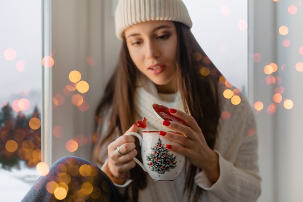 Young attractive woman in stylish white knitted sweater, scarf and hat sitting at home on windowsill at Christmas holding cup drinking hot tea, winter forest background view, lights bokeh
