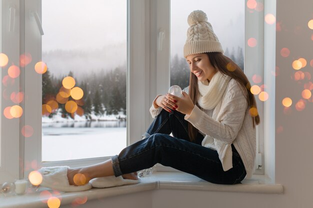 Young attractive woman in stylish white knitted sweater, scarf and hat sitting at home on windowsill at Christmas holding glass snow ball present decoration, winter forest view, lights bokeh