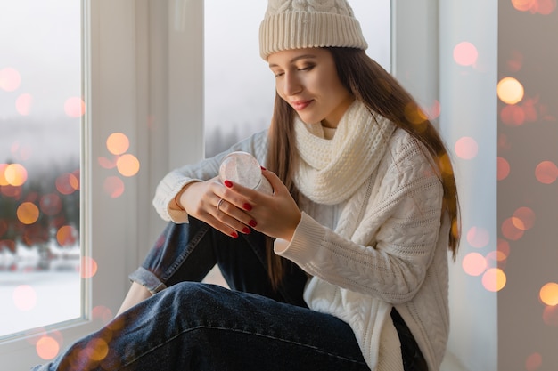 スタイリッシュな白いニットのセーター、スカーフ、帽子をかぶった若い魅力的な女性がクリスマスに窓辺に座ってガラスの雪玉プレゼントの装飾、冬の森の景色、ライトボケを保持しています