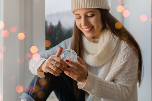 Молодая привлекательная женщина в стильном белом вязаном свитере, шарфе и шляпе сидит дома на подоконнике на Рождество, держа в руках стеклянный снежный шар, настоящее украшение, вид на зимний лес, огни боке