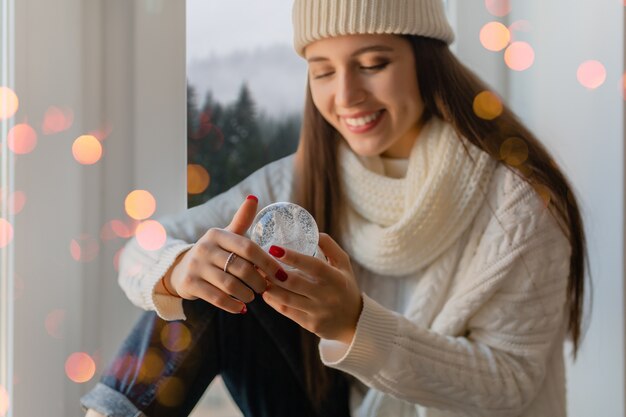 Young attractive woman in stylish white knitted sweater, scarf and hat sitting at home on windowsill at Christmas holding glass snow ball present decoration, winter forest view, lights bokeh