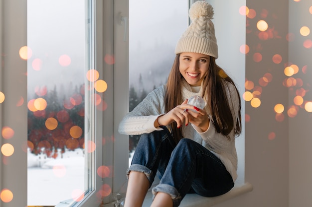 Young attractive woman in stylish white knitted sweater, scarf and hat sitting at home on windowsill at Christmas holding glass snow ball present decoration, winter forest view, lights bokeh