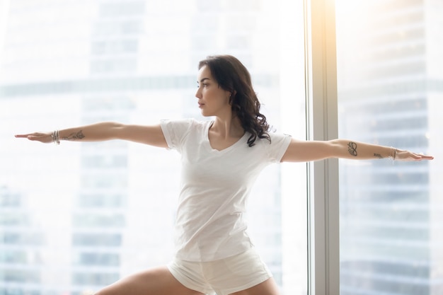 Young attractive woman in Warrior Two pose, against the window