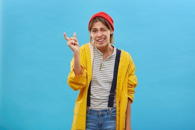 Young attractive woman wearing red hat, yellow jacket and jean overalls showing something very little with hands while gesturing. Fisherwoman demonstrating size of fish