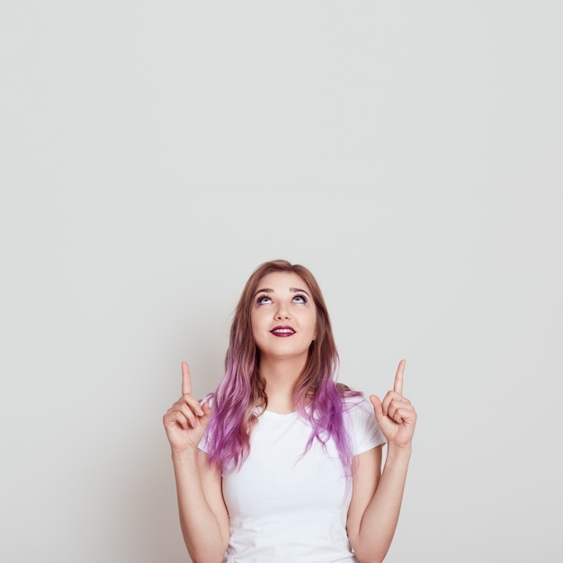 Молодая привлекательная женщина в белой повседневной футболке смотрит и указывает вверх обоими указательными пальцами, копирует пространство для рекламы или рекламного текста, позирует изолированно над серой стеной.