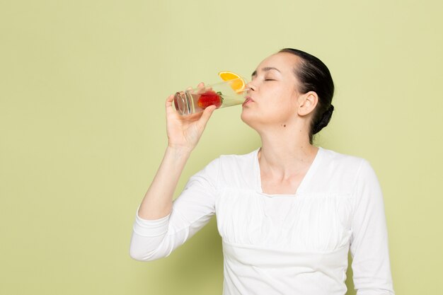 緑の背景にイチゴの中の水でガラスを保持している白いシャツの若い魅力的な女性