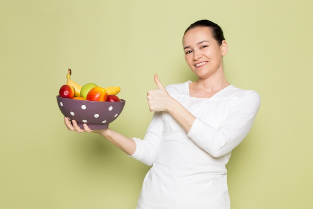 Giovane donna attraente in camicia bianca che sorride e che mostra la ciotola di frutta