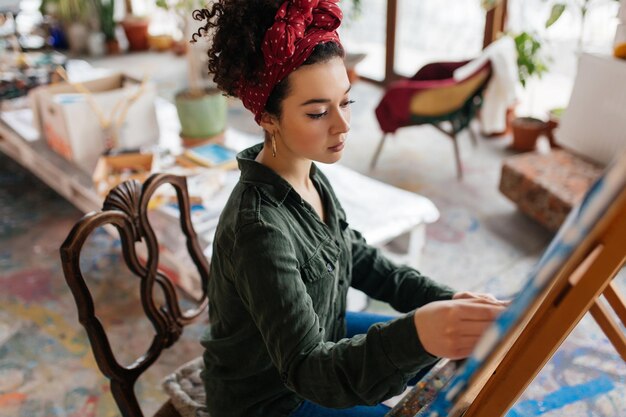 Молодая привлекательная женщина с темными вьющимися волосами сидит на стуле и мечтательно рисует картину на холсте в современной уютной художественной мастерской с большими окнами