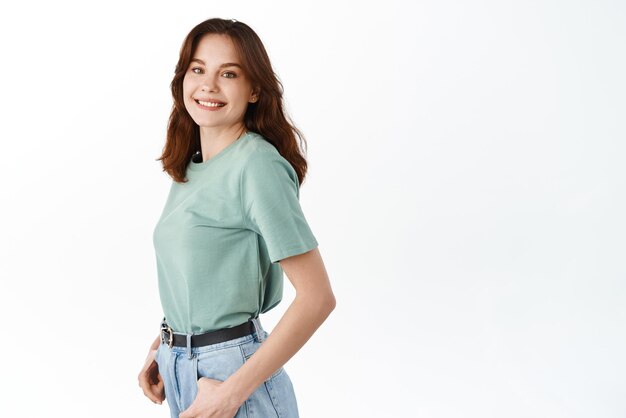 Young attractive woman with natural harido and make up smiling broadly at camera turn head at camera while standing in profile against white background