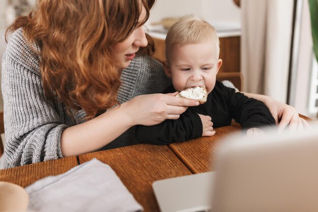 Young attractive woman with red hair in knitted sweater dreamily feeding her little son watching cartoons on laptop Mom spending time with baby in cozy kitchen at home